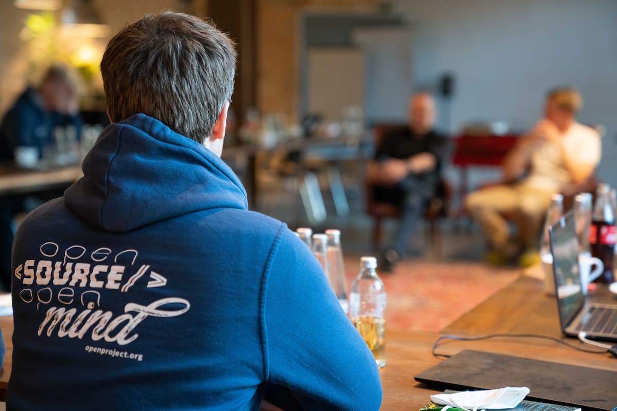 A person wearing a dark blue hooded sweatshirt has the OpenProject logo prominently displayed on the back. The logo features stylized text and a graphic design element in white.