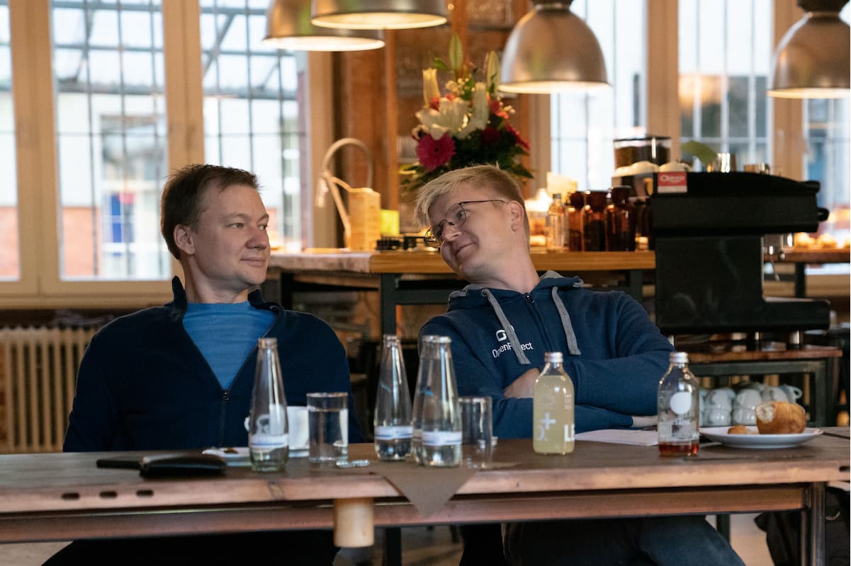 Two men seated at a wooden dining table with food and drinks, engaged in conversation.